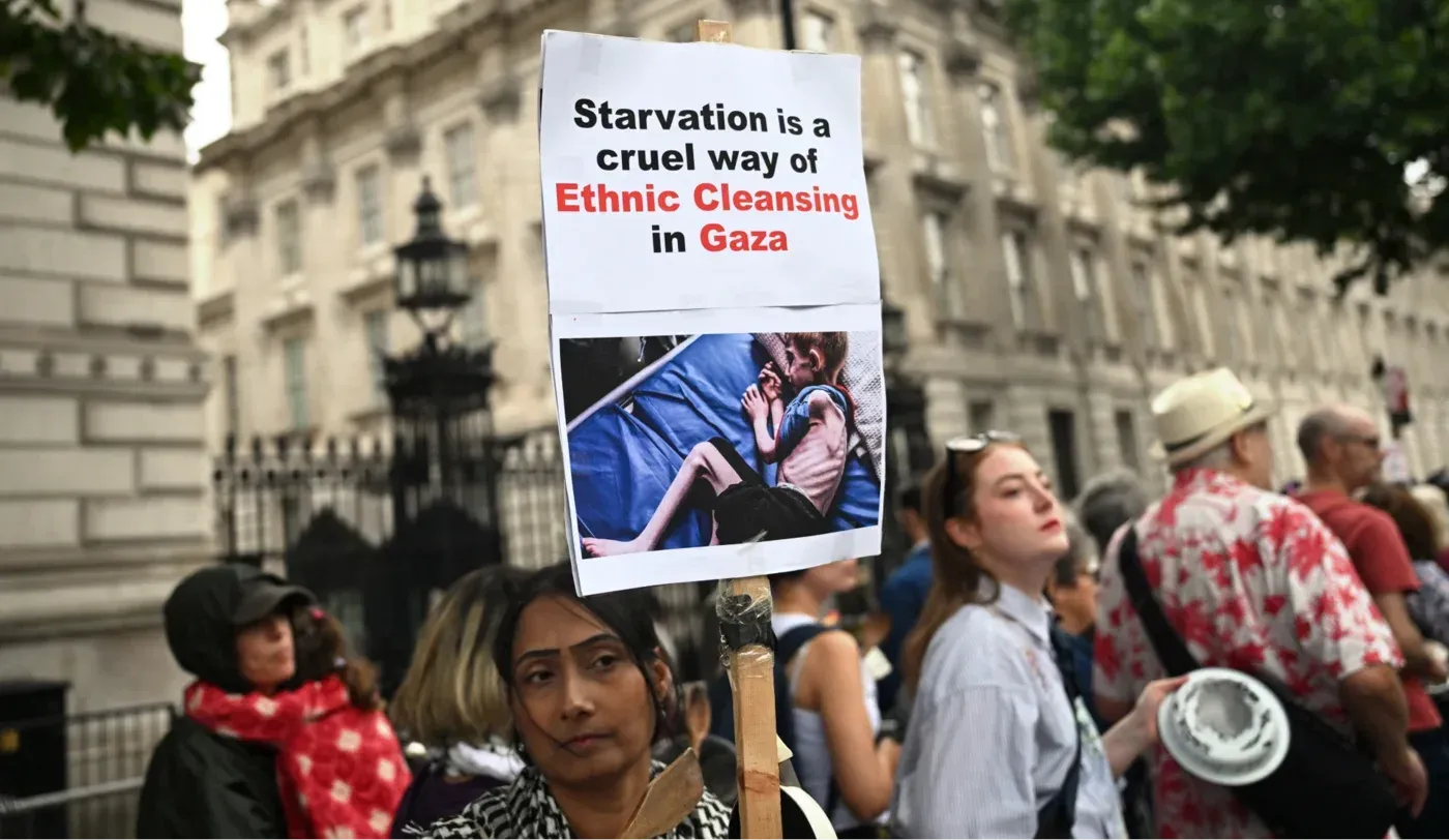 لافتة في مظاهرة تشير إلى أن التجويع هو وسيلة قاسية للتطهير العرقي في غزة، مع صورة لطفل يتلقى الطعام.