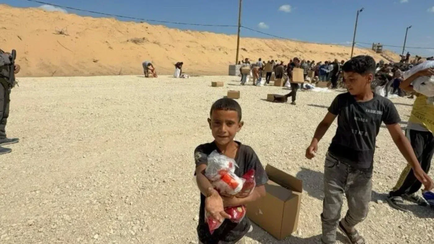 طفل فلسطيني يحمل مساعدات غذائية في منطقة توزيع، مع وجود مجموعة من الأشخاص في الخلفية، يعكس الوضع الإنساني الصعب في غزة.