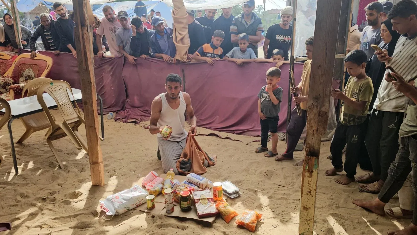 مشهد لفلسطيني يجلس على الأرض محاطًا بالحشود، بينما يعرض المساعدات الغذائية في موقع توزيع تحت خيمة في غزة.