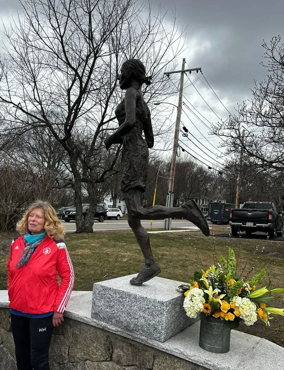 تمثال برونزي يُظهر Bobbi Gibb، أول امرأة تشارك في ماراثون بوسطن، بجانب زهور ملونة، مع خلفية من الأشجار والسماء الرمادية.