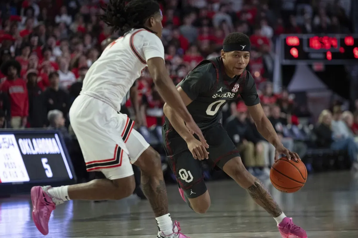 لاعب كرة سلة من فريق أوكلاهوما يت dribble الكرة بينما يواجه مدافعًا، مع جمهور متحمس في الخلفية.