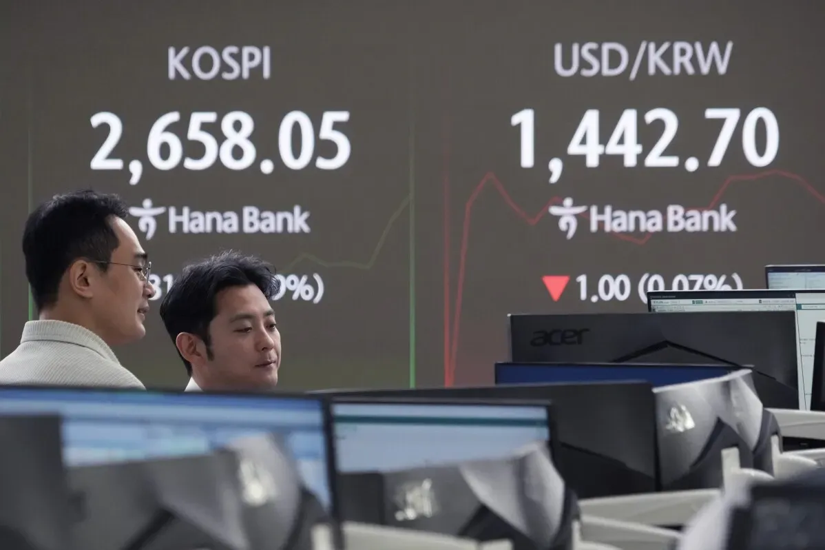 شخصان يتابعان مؤشرات الأسهم في بورصة كورية، مع عرض لمؤشر KOSPI وسعر الدولار مقابل الوون الكوري على الشاشة.