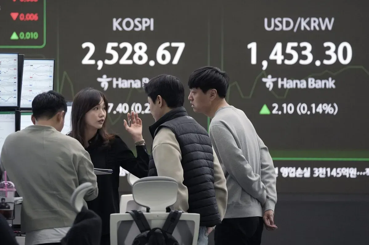 مجموعة من الأشخاص يتحدثون في مركز مالي، مع عرض لمؤشر KOSPI وأسعار العملات على الشاشة خلفهم.