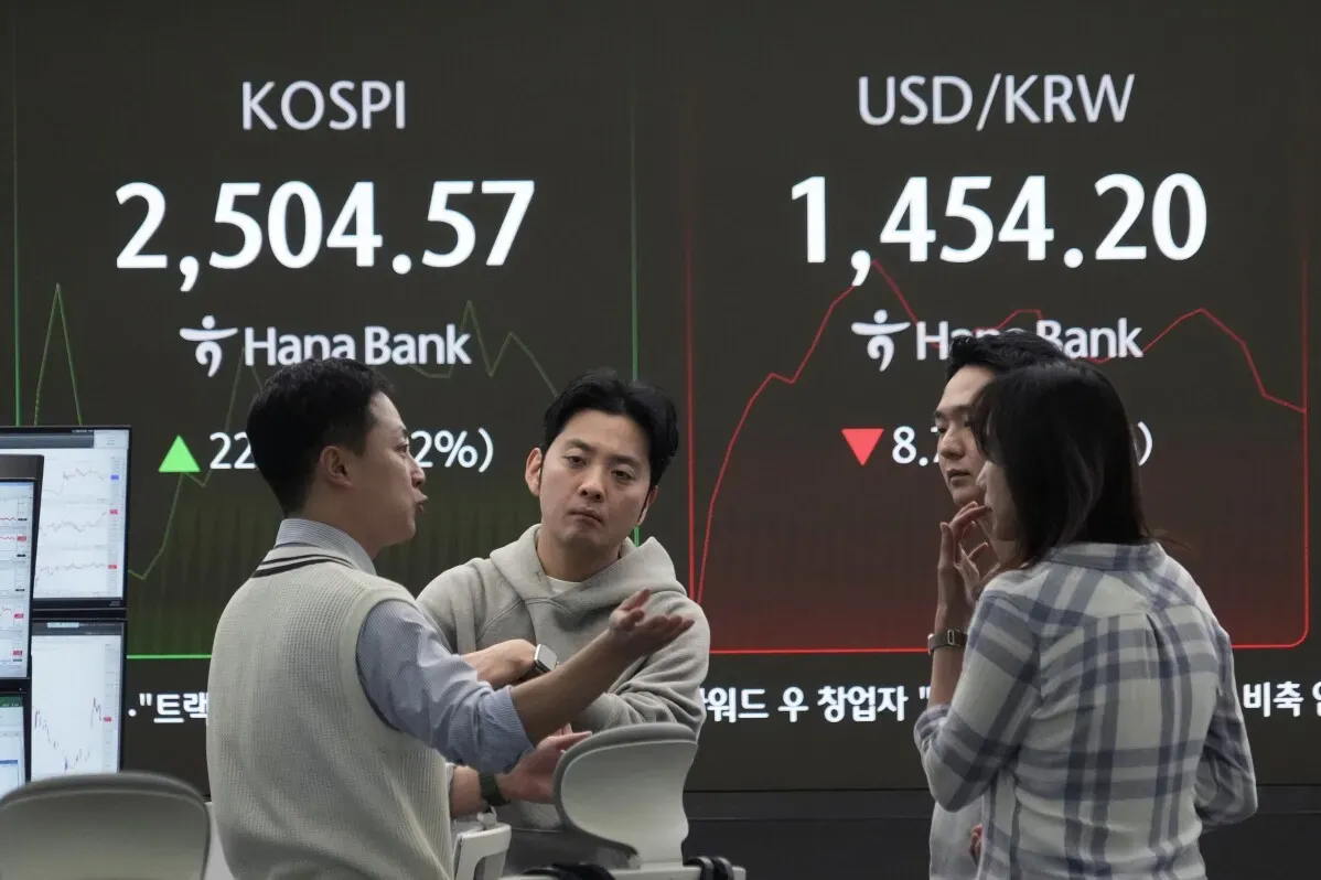 مستثمرون يتناقشون في بورصة كوسبي الكورية الجنوبية، مع عرض أسعار الأسهم والدولار مقابل الوون على الشاشة الخلفية.