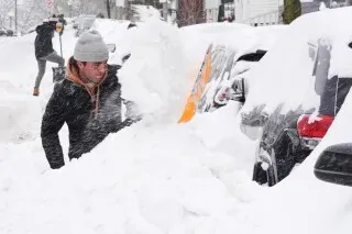 رجل يعمل على إزالة الثلوج من سيارة مغطاة بالكامل في منطقة تعرضت لعاصفة جليدية، مع تساقط الثلوج الكثيفة حوله.