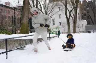 امرأة تسحب طفلًا في زلاجة عبر الثلوج العميقة، تعكس أجواء الشتاء القاسي بعد العاصفة الجليدية.