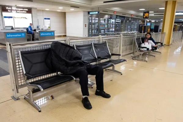 شخص يرتدي سترة سوداء يجلس بلا حراك على مقعد في مطار، بينما تجلس امرأة أخرى على مقعد قريب، مما يعكس الازدحام والاضطرابات في السفر الجوي.