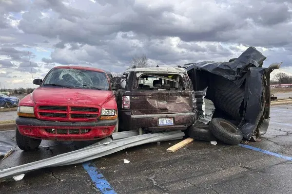 تظهر الصورة سيارات متضررة في موقف سيارات بعد عواصف قوية في ميشيغان، مع وجود حطام معدني حولها، مما يعكس تأثير الطقس القاسي.