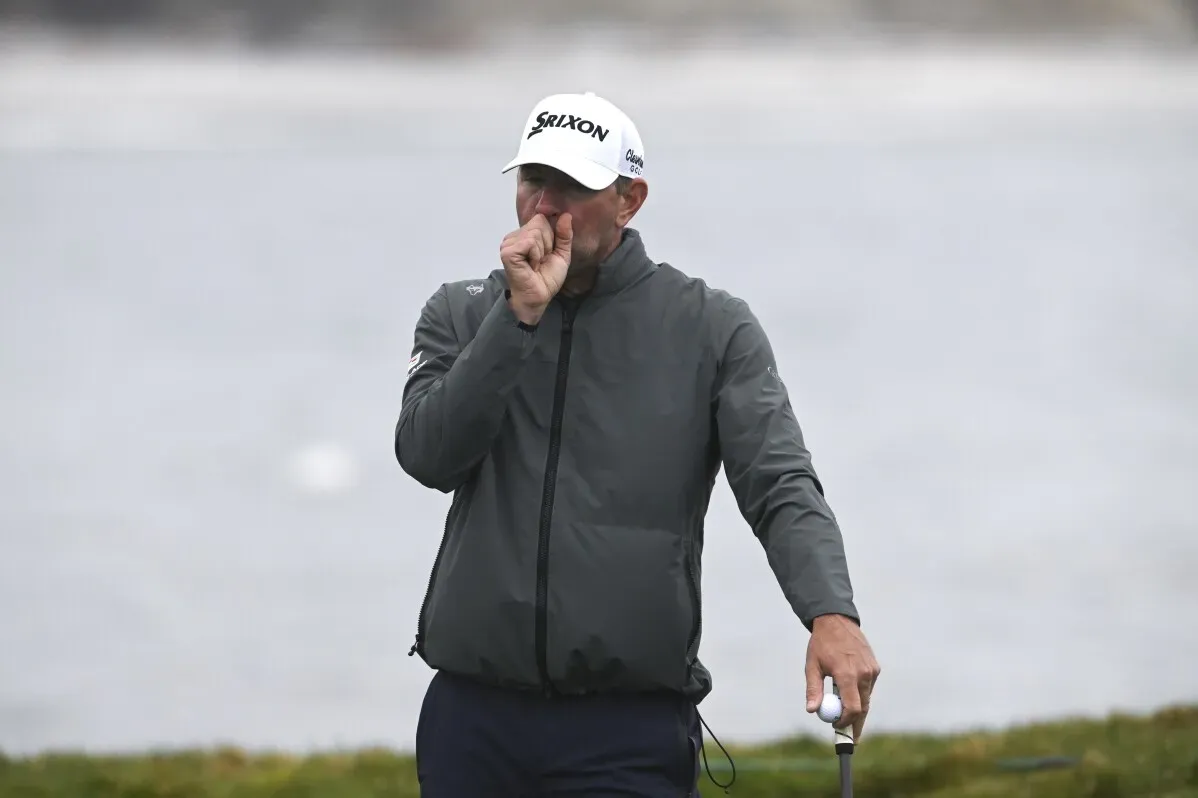لاعب غولف يرتدي سترة رمادية وقبعة بيضاء، يبدو متفكراً أثناء جولته في بطولة AT&T Pebble Beach Pro-Am تحت ظروف جوية عاصفة.
