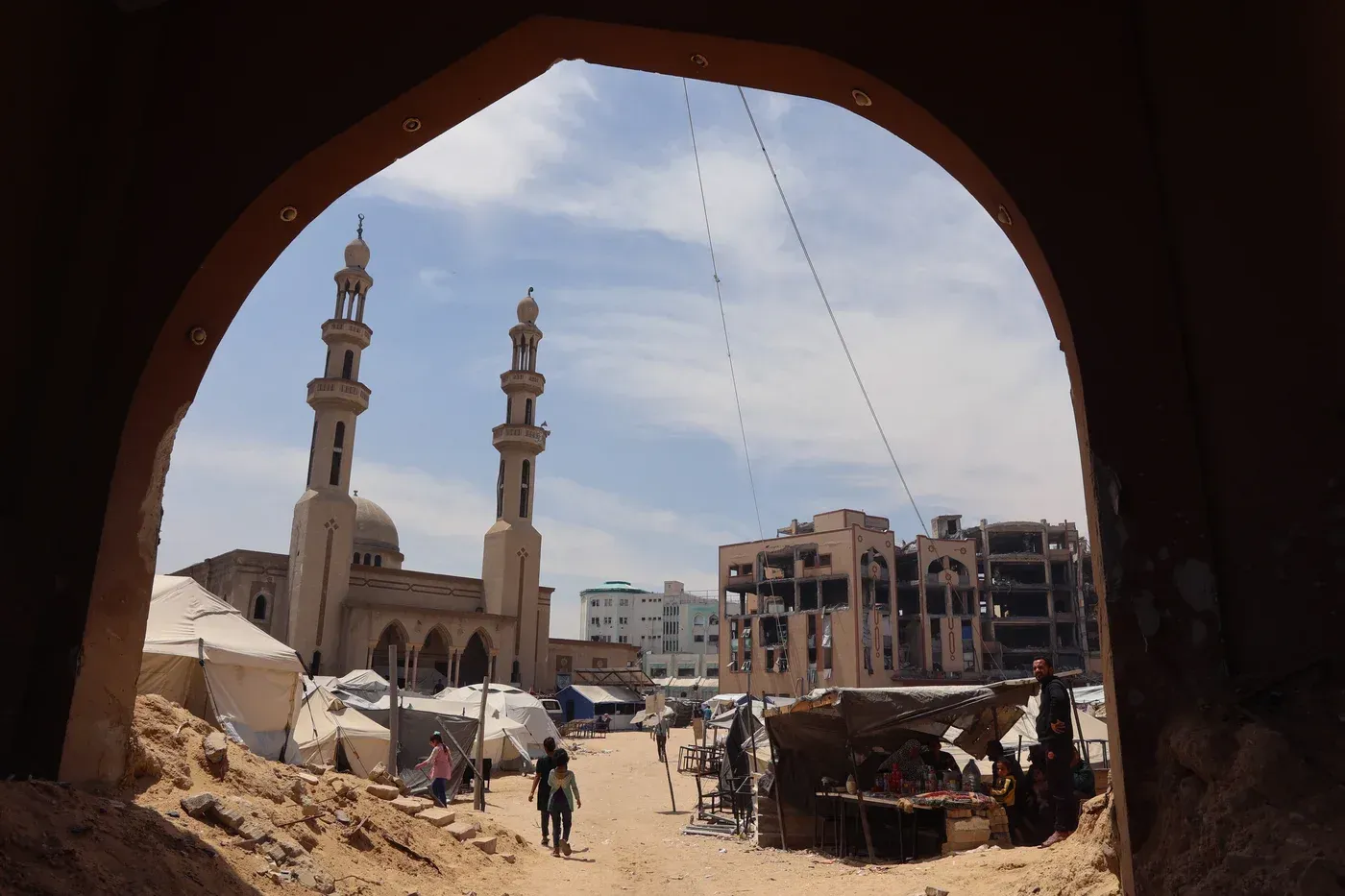 ساحة في غزة تضم خيامًا للنازحين، مع مسجد في الخلفية ومباني مدمرة، تعكس الأثر المدمر للصراع المستمر.