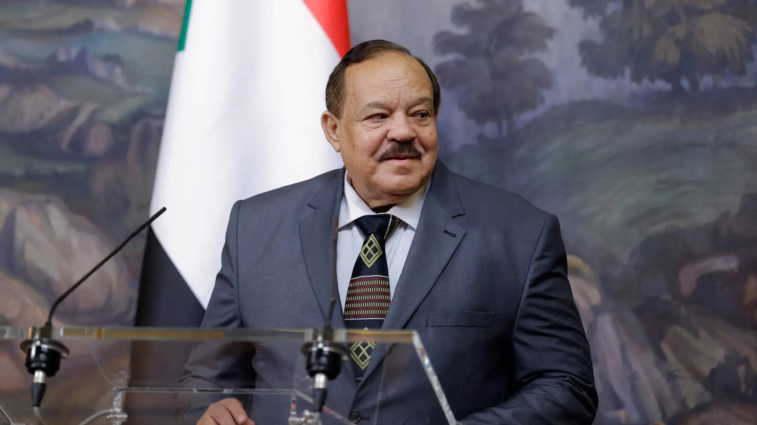 وزير الخارجية السوداني علي يوسف الشريف يتحدث في مؤتمر صحفي، مع العلم السوداني في الخلفية، وسط أجواء دبلوماسية.