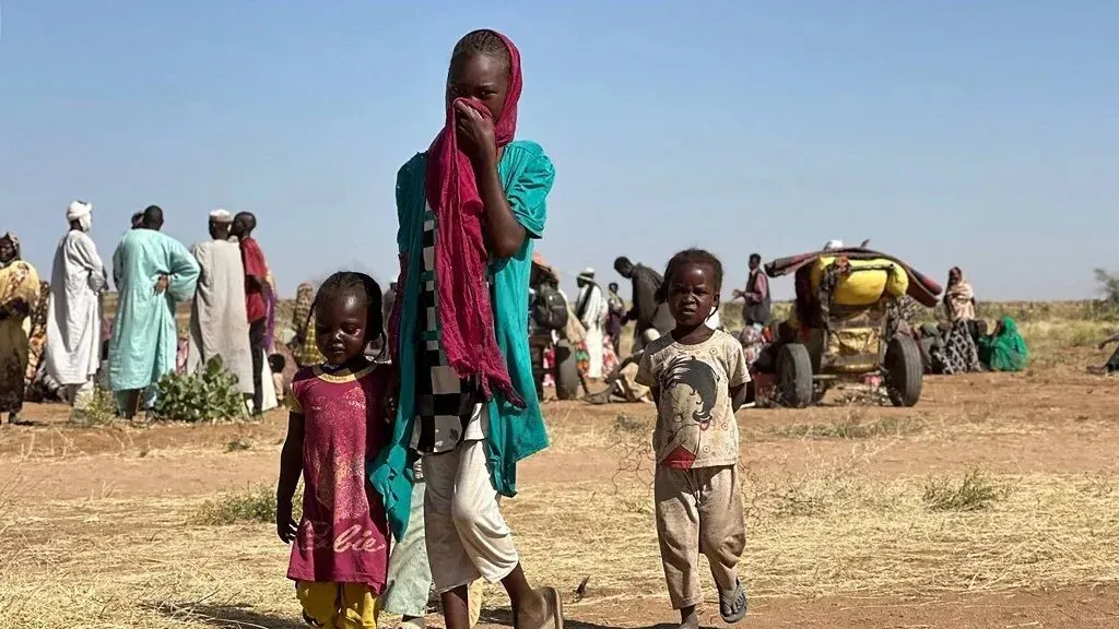 أطفال نازحون في دارفور يقفون في مقدمة الصورة بينما يظهر خلفهم آخرون في منطقة قاحلة، مما يعكس آثار النزاع والأزمة الإنسانية.