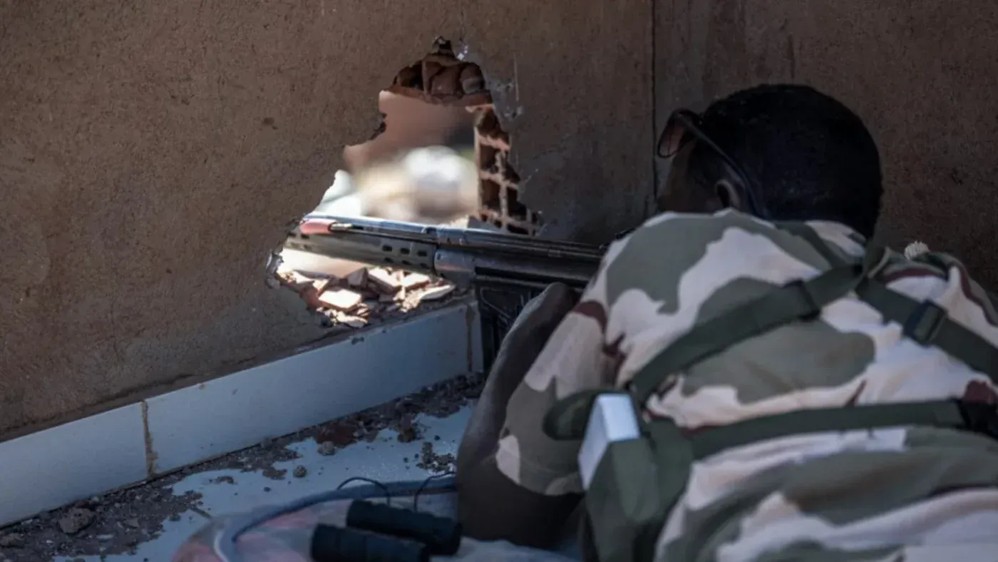 مقاتل يرتدي زيًا عسكريًا يستعد لإطلاق النار من خلال ثقب في الجدار، مما يعكس حالة العنف المستمرة في السودان وتأثير الحرب على الصحافة.