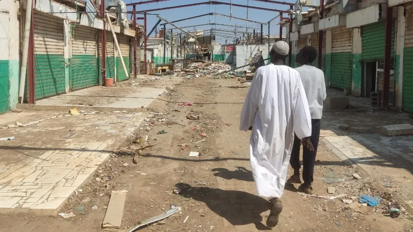 شارع مدمّر في قرية سودانية، يظهر فيه رجلان يسيران وسط الأنقاض والمخلفات، مما يعكس آثار النزاع المستمر في البلاد.