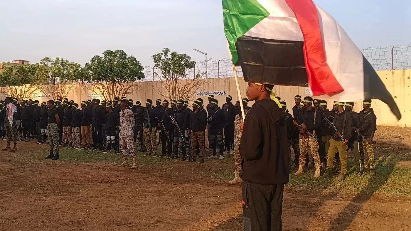 مجموعة من المقاتلين يرتدون زيًا موحدًا، يتجمعون في ساحة، بينما يحمل أحدهم علم السودان، في سياق تجنيدهم للقتال ضد قوات الدعم السريع.