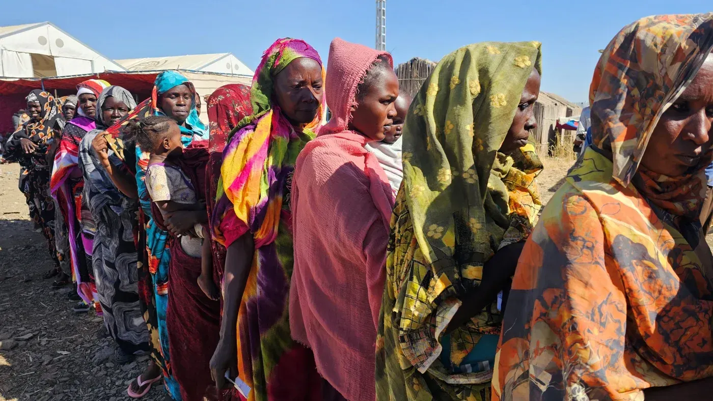 نساء سودانيات يرتدين أزياء ملونة، ينتظرن في صف للحصول على المساعدات الإنسانية، مما يعكس تأثير النزاع على المدنيين.