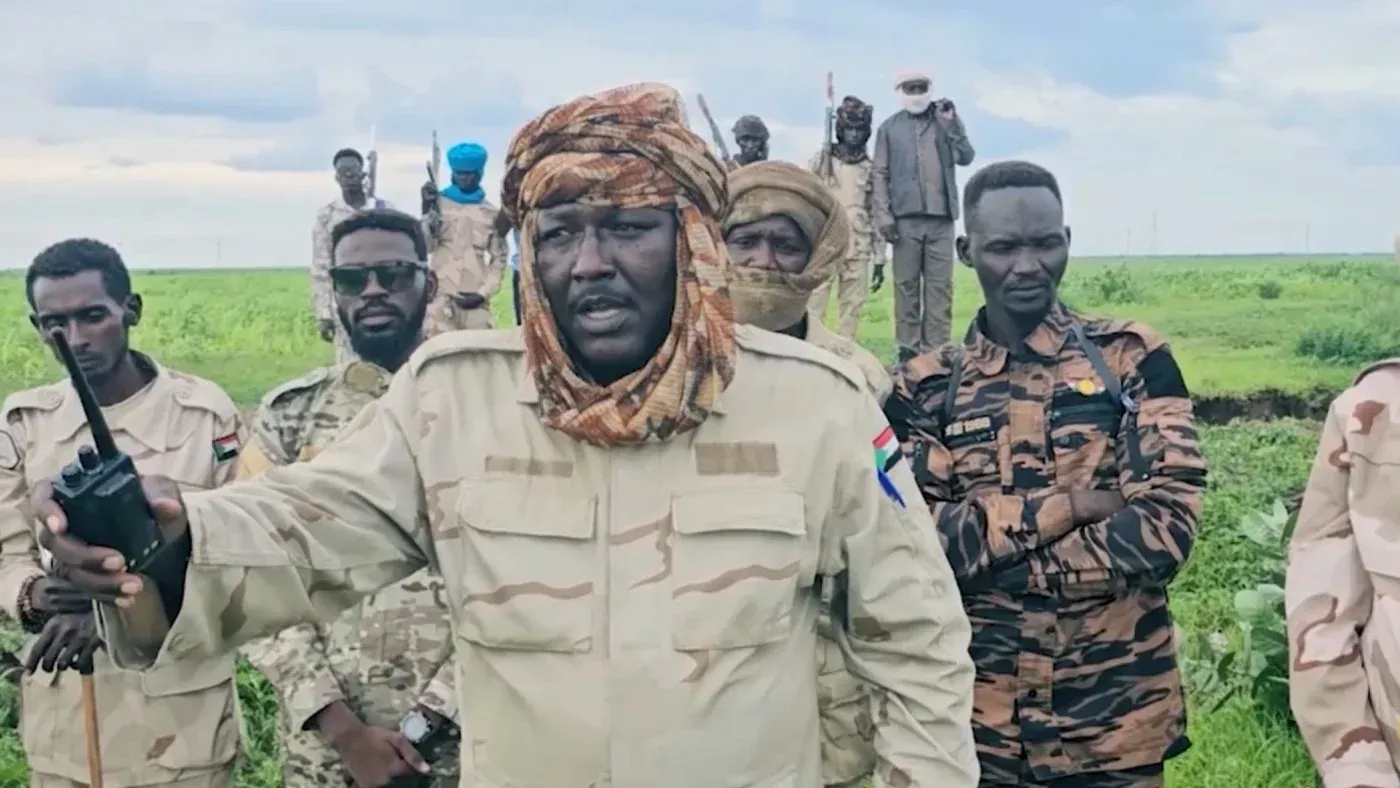 قائد من قوات الدعم السريع يتحدث عبر جهاز لاسلكي، محاط بمقاتلين في منطقة خضراء، في سياق الهجوم على القوات المسلحة السودانية.