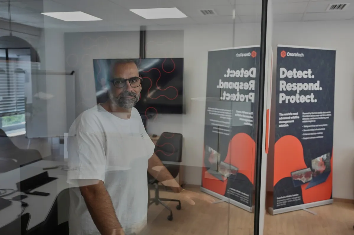 رجل يرتدي نظارات يقف داخل مكتب حديث، خلفه لافتات لشركة OroraTech تتحدث عن رصد الحرائق والاستجابة لها، مما يعكس الابتكار في مكافحة الحرائق.
