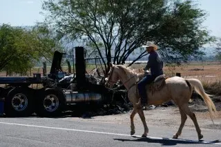 رجل على حصان أبيض يسير بجانب شاحنة محترقة في منطقة قاحلة، مما يعكس تأثير العنف المرتبط بعصابات المخدرات في المكسيك.