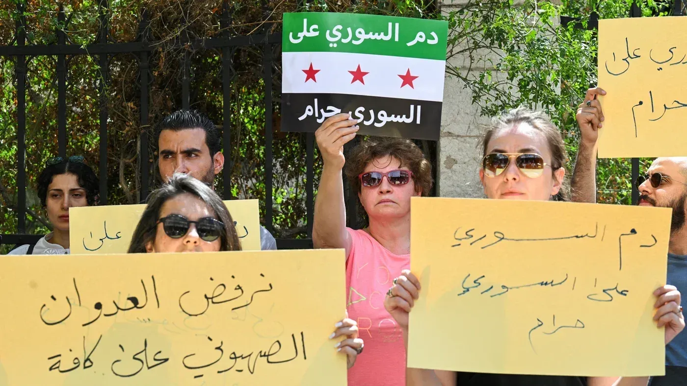محتجون يحملون لافتات تدعو إلى دعم سوريا ورفض العدوان الإسرائيلي، مع وجود علم الثورة السورية في الخلفية.