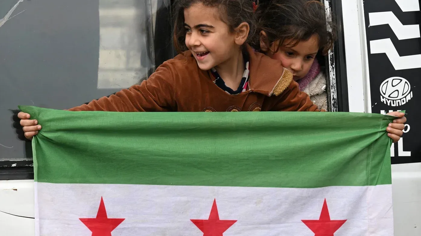 طفلتان تحملان علم سوريا القديم، مع ابتسامة على وجهيهما، تعكسان الأمل في إعادة بناء البلاد بعد الصراع.