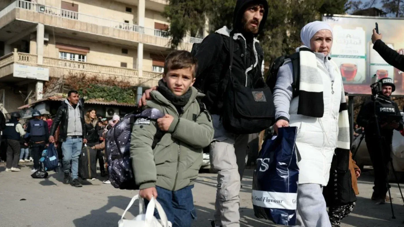 عائلة سورية تتجول في منطقة حضرية، حيث يحمل طفل حقيبة ظهر، بينما يحمل البالغون أكياسًا، مما يعكس آثار النزاع المستمر في البلاد.