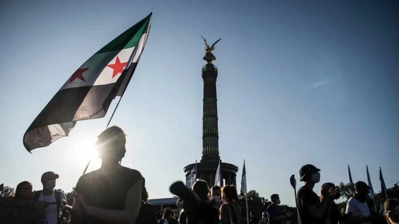علم سوري يحمل رموز الثورة، يتوسط مظاهرة تحت أشعة الشمس، مع عمود وقاعدة معروفين في الخلفية. تشييع لمشاعر الأمل والقلق حول العودة.