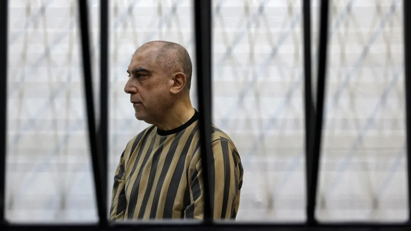 عاطف نجيب، الرئيس السابق لجهاز الأمن، يجلس في قفص المحكمة مرتديًا بدلة سجن مخططة، في محاكمة تتعلق بجرائم ضد الشعب السوري.