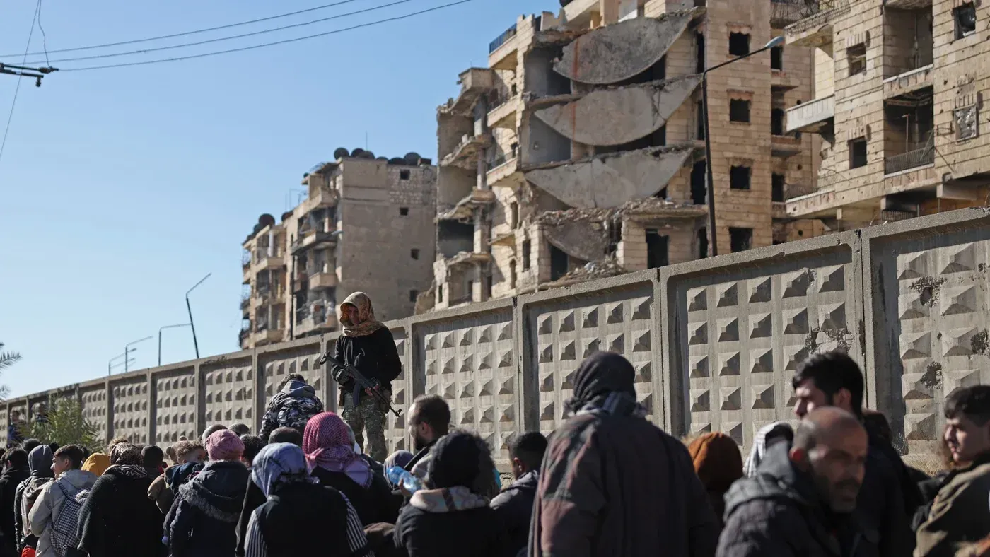 مشهد لمدنيين نازحين في حي الشيخ مقصود بحلب، مع وجود مبانٍ مدمرة في الخلفية، وسط تصاعد العنف بين القوات السورية والأكراد.