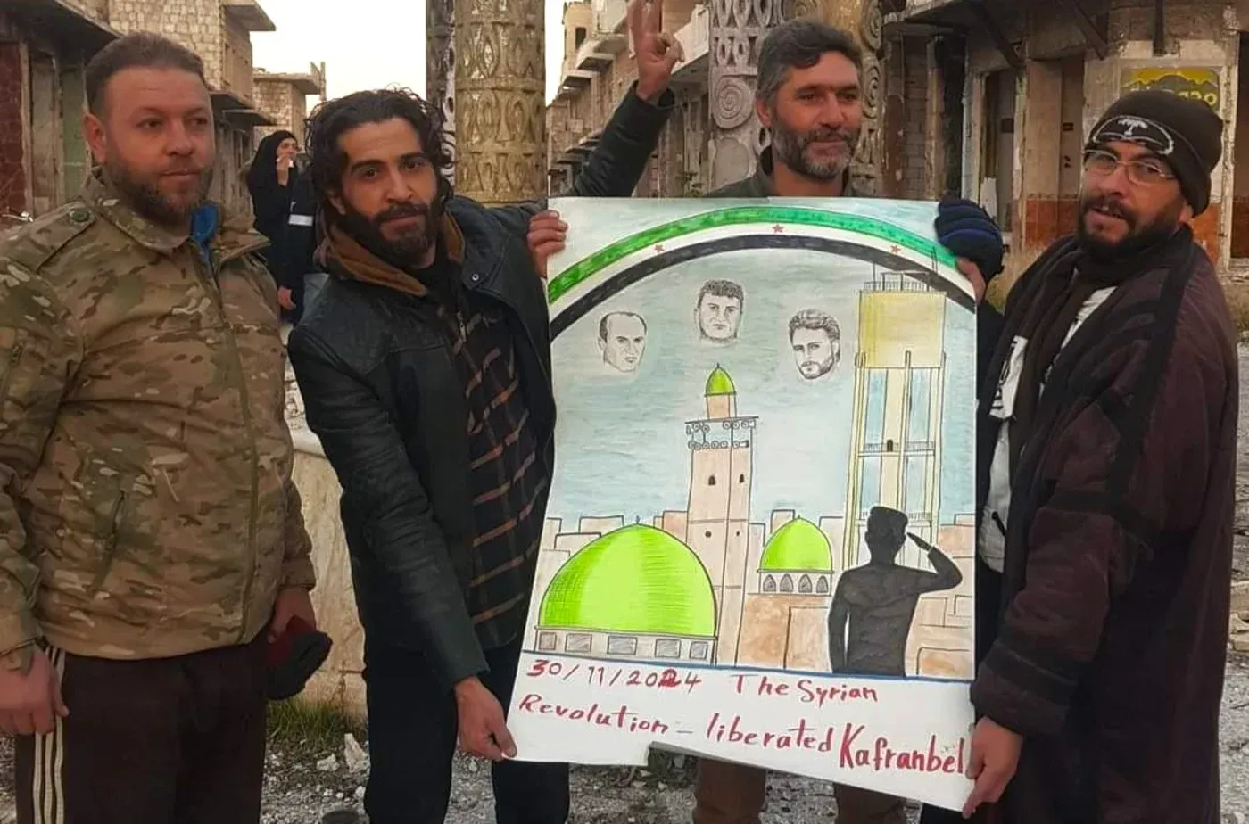 ناشطون في كفرنبل يحملون لوحة فنية تحت قوس قزح، تبرز صور رائد فارس ورفاقه، تعبيرًا عن دعمهم للثورة السورية.