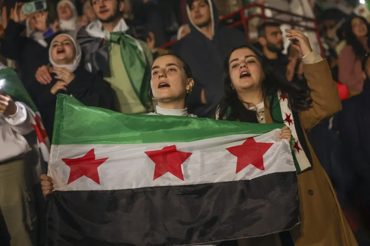 نساء يحملن علم الثورة السورية ويغنين بحماس خلال حفل لوصفي المعصراني في دمشق، مع تفاعل الجمهور وإضاءة الهواتف.