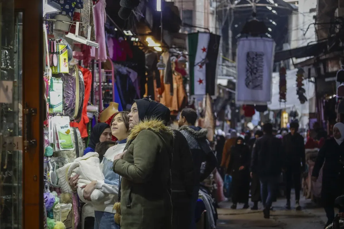 نساء يتجولن في سوق قديم مزدحم بدمشق، يطلعن على السلع المعروضة بينما يرفرف العلم السوري في الخلفية، مما يعكس الأجواء الاحتفالية.