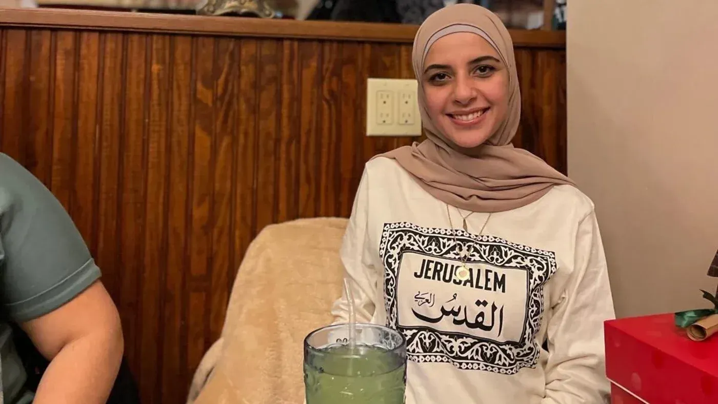 لقاء كوردية، شابة ترتدي حجابًا، تجلس في مطعم مع مشروب أمامها، تحمل قميصًا مكتوبًا عليه "القدس".
