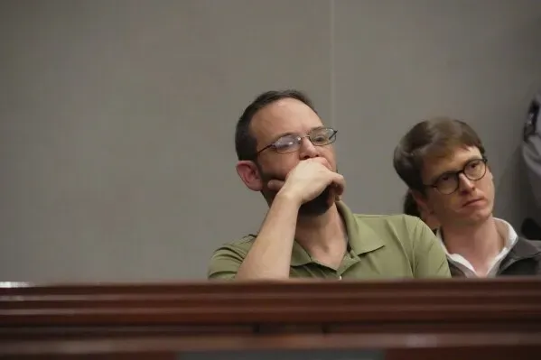 شخصان يجلسان في قاعة المحكمة، أحدهما يرتدي نظارات ويبدو عليه التأمل، بينما الآخر ينظر إلى الأمام بجدية، مما يعكس أجواء المحاكمة المتعلقة بإطلاق النار الجماعي.