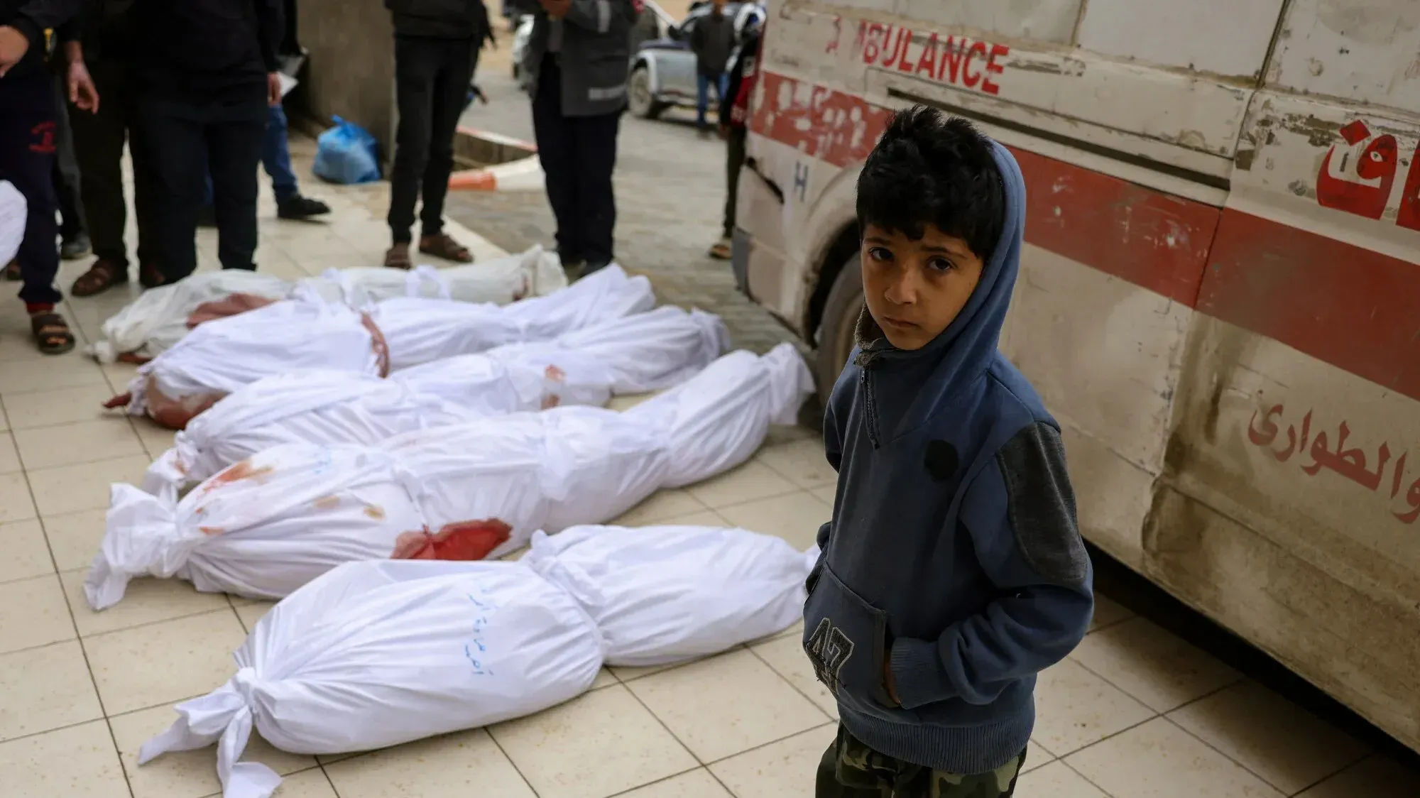 طفل فلسطيني يقف بجانب جثث مغطاة في مستشفى بغزة بعد غارات جوية، حيث قُتل أكثر من 400 شخص، بينهم أطفال ونساء، خلال الهجمات.