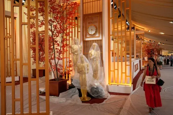 ديكور حفل توزيع جوائز الأوسكار مع تماثيل مغطاة وديكورات زهرية، بينما تسير امرأة تحمل لافتة.