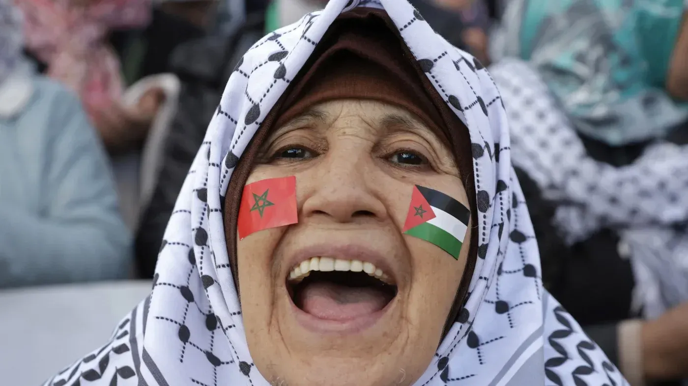 امرأة مسنّة ترتدي حجابًا مميزًا، تعبر عن دعمها لفلسطين والمغرب من خلال وضع علميهما على وجهها، خلال مظاهرة حاشدة.