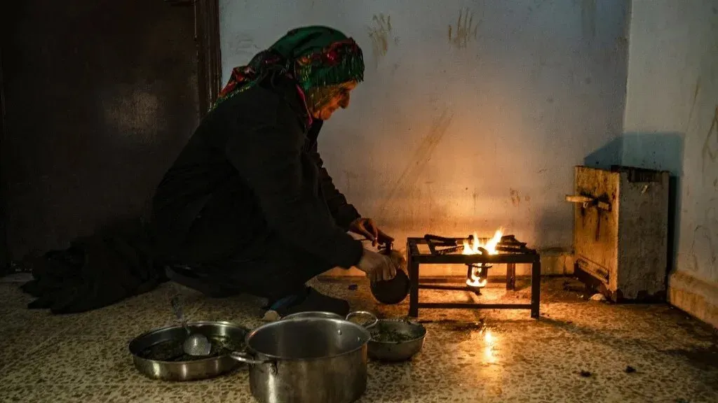 امرأة مسنّة تحضر الطعام على موقد بسيط وسط غرفة مظلمة، مع وجود أواني معدنية حولها، تعكس الظروف المعيشية الصعبة في شمال شرق سوريا.