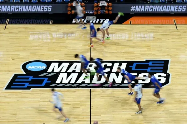 لاعبو كرة السلة يتنافسون في مباراة NCAA على أرضية ملعب تحمل شعار "March Madness"، مع كرة سلة في الخلفية.