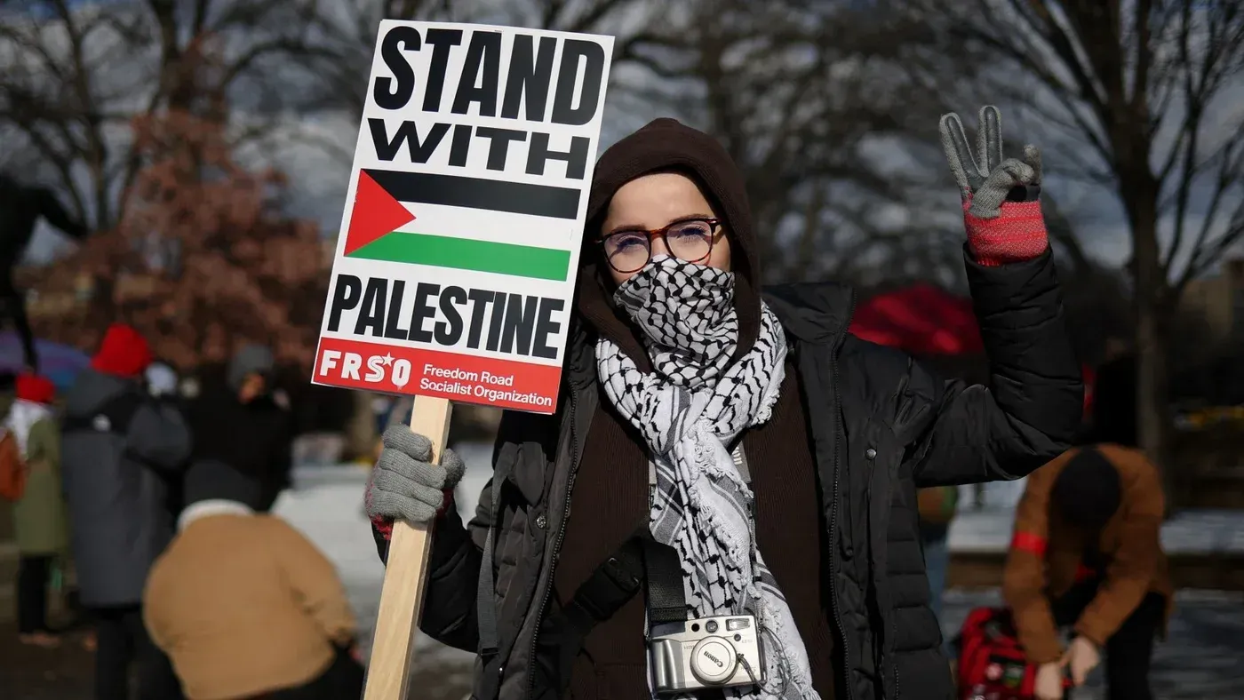 شخص يحمل لافتة مكتوب عليها "أقف مع فلسطين" خلال تظاهرة، مع خلفية تضم عددًا من المشاركين الآخرين في الفعالية.