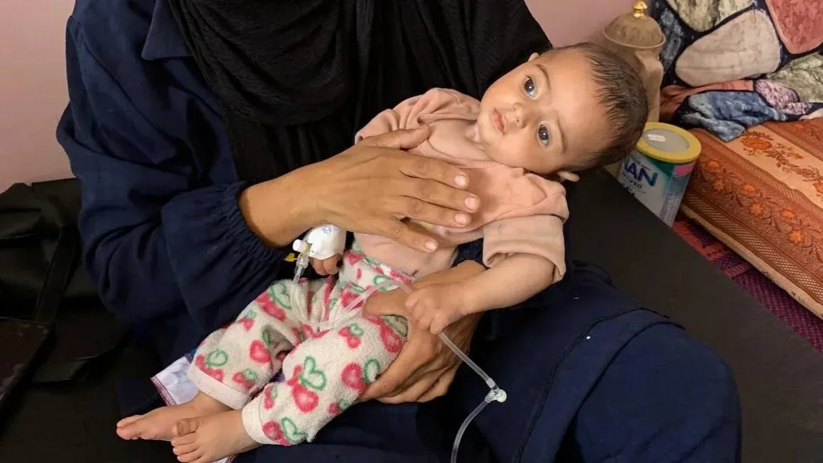 رضيع ضعيف يعاني من التهاب السحايا، يُحمل بين ذراعي والدته في غرفة بسيطة، مع أنبوب طبي يربط بينهما، وسط ظروف إنسانية صعبة في غزة.
