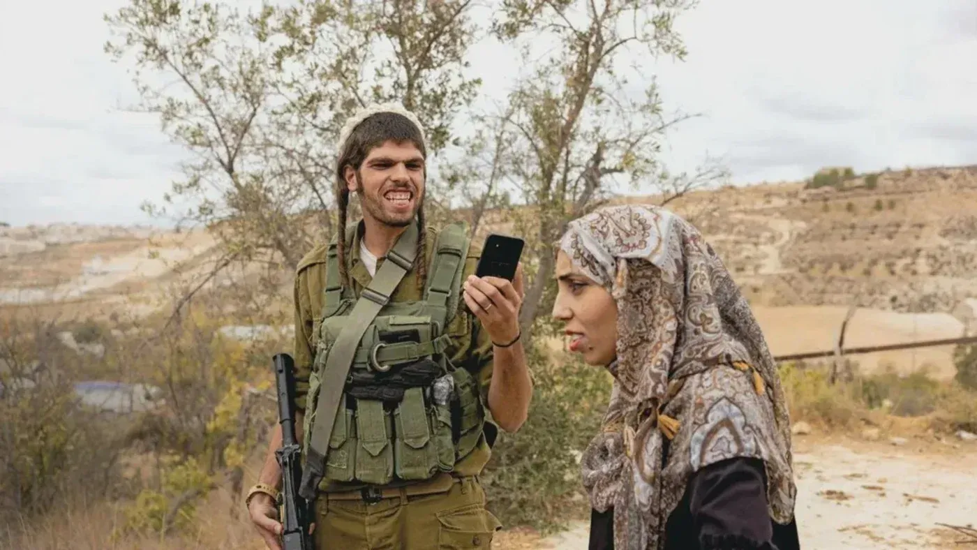 مستوطن إسرائيلي مسلح يضحك ويصور معاذ أبو الرب بهاتفه، بينما تظهر معاذ في حالة تحدٍ خلال احتجاج ضد الاستيطان.