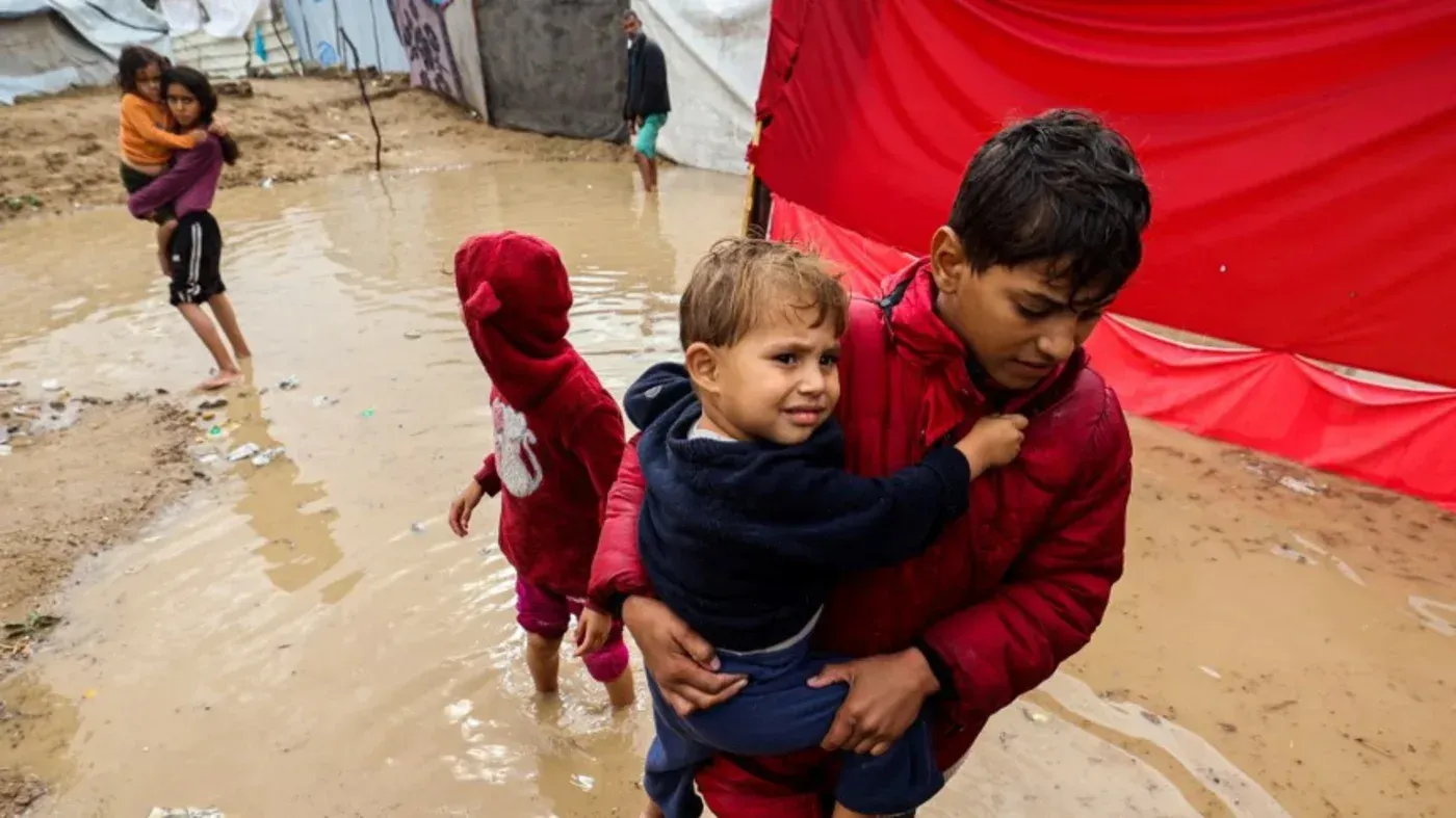 أطفال في مخيم للاجئين وسط مياه الأمطار، يعانون من ظروف قاسية في غزة، حيث تشتد أزمة المساعدات الإنسانية.