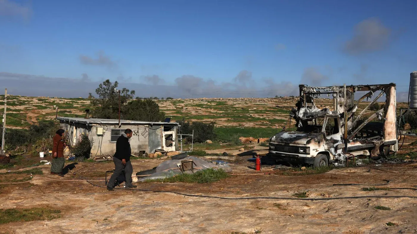 مشهد لقرية فلسطينية تظهر فيها قافلتان بيضاوتان متوقفتان بجانب منزل متضرر، مع شخصين يسيران في المنطقة. تعكس الصورة التوترات المستمرة في الضفة الغربية.