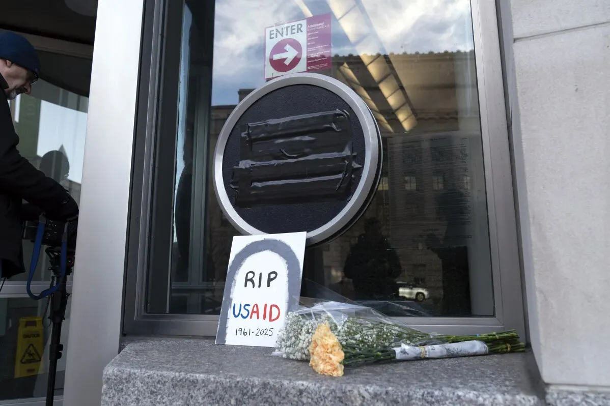 تظهر الصورة لافتة مكتوب عليها "RIP USAID 1961-2025" مع زهور موضوعة أمام نافذة مغلقة، تعبيرًا عن الاحتجاج على تقليص وكالة المساعدات الأمريكية.