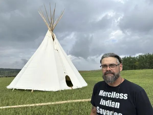 رجل يقف أمام خيمة تقليدية في منطقة طبيعية، مرتديًا قميصًا يحمل عبارة "Merciless Indian Savages"، تعبيرًا عن هويته الثقافية.