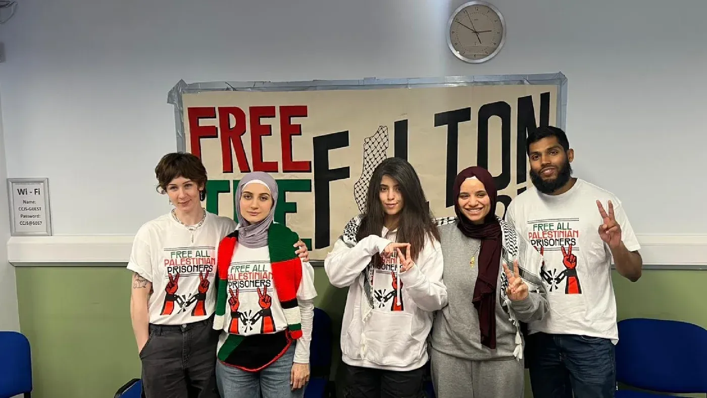 مجموعة من الأشخاص يرتدون قمصانًا تحمل شعارات تطالب بالإفراج عن جميع السجناء الفلسطينيين، يقفون أمام لافتة كبيرة تحمل نفس الرسالة.