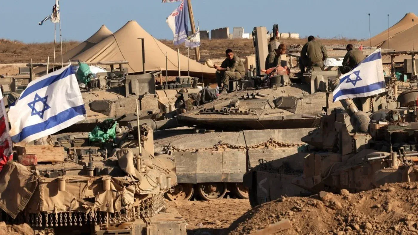 جنود إسرائيليون في قاعدة عسكرية، مع دبابات تحمل العلم الإسرائيلي، يبرز دورهم في العمليات العسكرية في غزة.