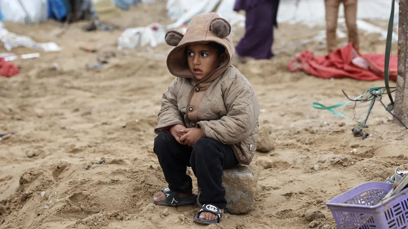 طفل فلسطيني يرتدي معطفًا ثقيلًا يجلس على الأرض في مخيم للنازحين في غزة، وسط ظروف قاسية ونقص في المأوى والتدفئة.