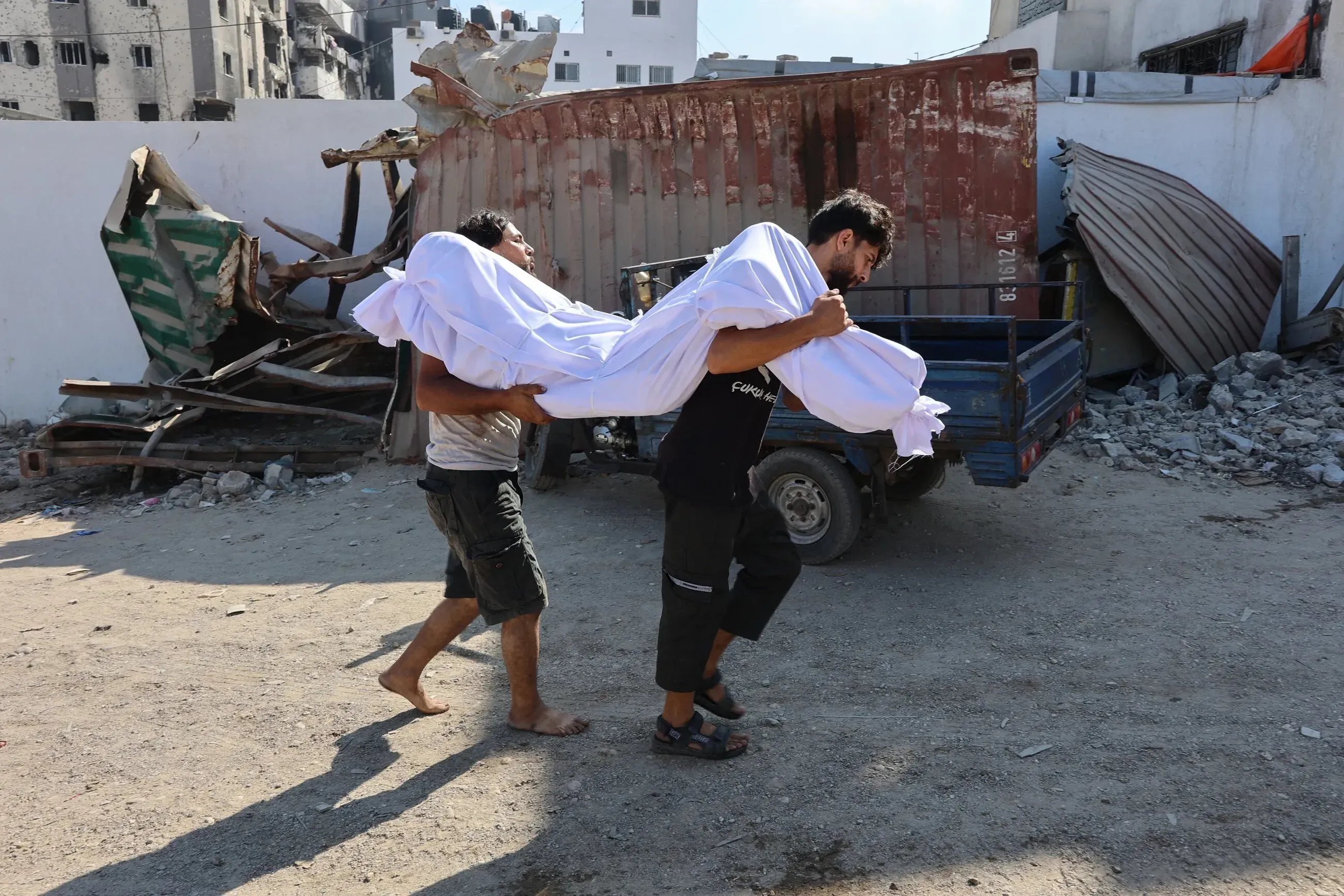 رجلان يحملان جثمانًا مغطى بملاءة بيضاء في غزة، وسط دمار خلفته الهجمات، مما يعكس الأزمة الإنسانية المتفاقمة في المنطقة.
